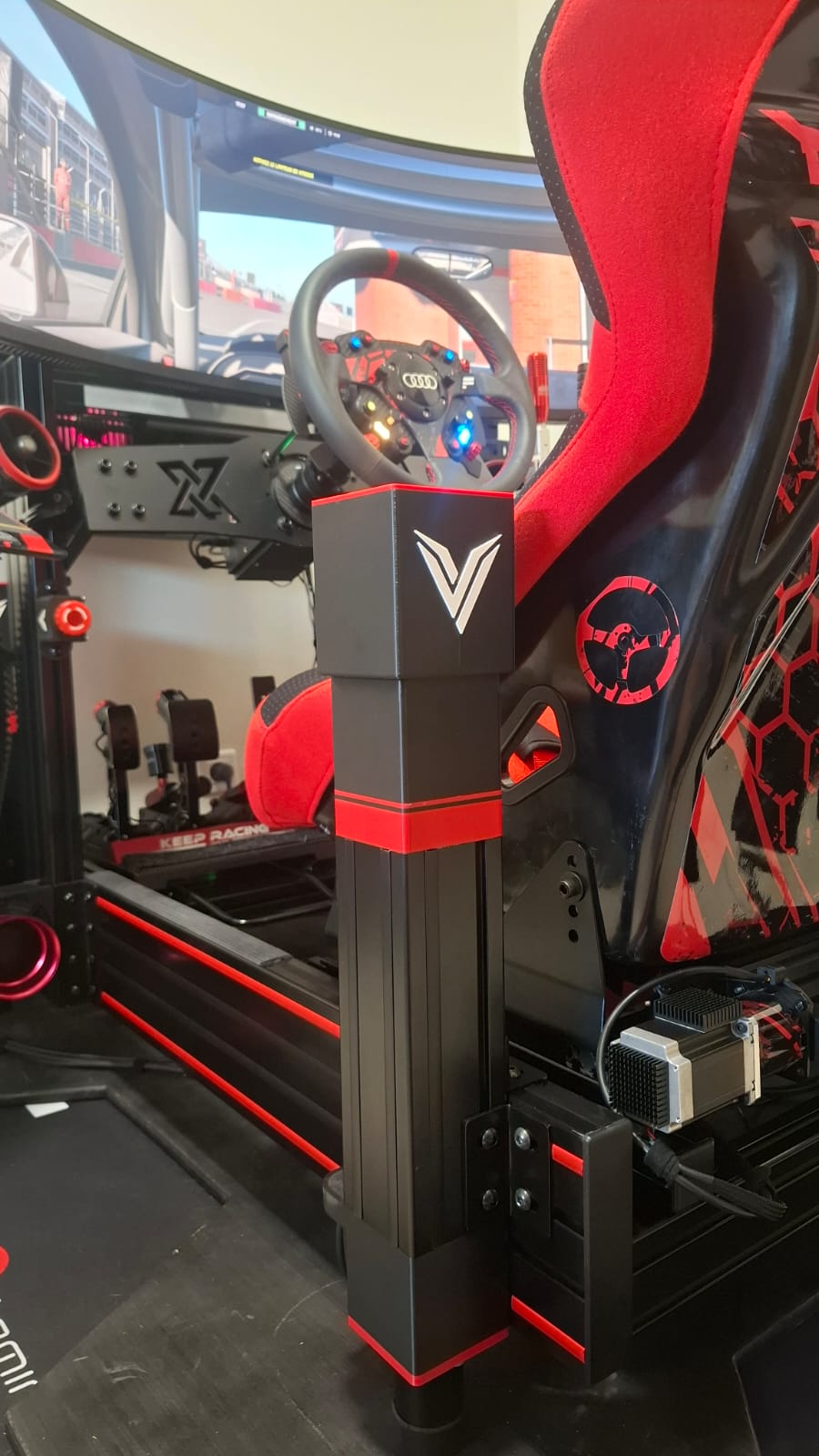 Vérin V-TRAX en finition noire et rouge, monté sur un simulateur de sim racing à côté d'un siège baquet
